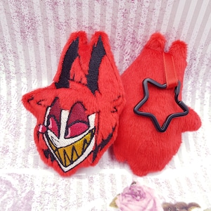 Könnte beinhalten: Roter Plüsch-Schlüsselanhänger mit einem Cartoon-Gesicht eines roten Dämons mit einem breiten Grinsen und gelben Zähnen. Der Schlüsselanhänger hat einen schwarzen sternförmigen Anhänger.