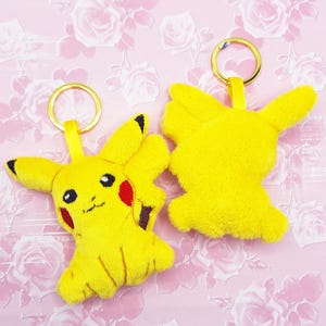 May include: Two yellow plush Pikachu keychains with black eyes, red cheeks, and black-tipped ears. Each keychain has a gold-colored ring and a yellow fabric loop. The keychains are on a pink floral background.