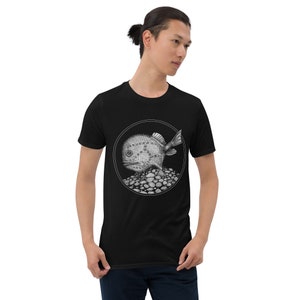 May include: Black t-shirt featuring a detailed, grayscale illustration of a round fish with fins and a textured body, set within a circular frame. The fish rests on a bed of small stones. The shirt is worn by a person.