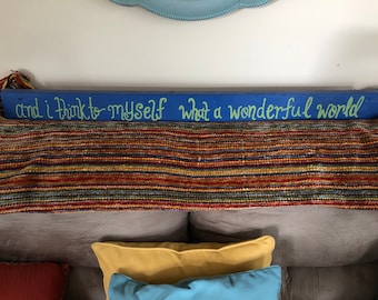 May include: A blue wooden sign with the text "and I think to myself what a wonderful world" written in green paint. The sign is hanging above a striped blanket.
