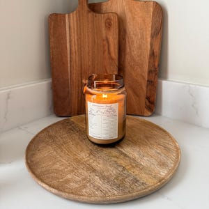 May include: A lit candle in a glass jar with a label sits on a wooden tray. Two wooden cutting boards stand behind the candle. The candle's flame glows, and the wooden items have a warm, natural color.