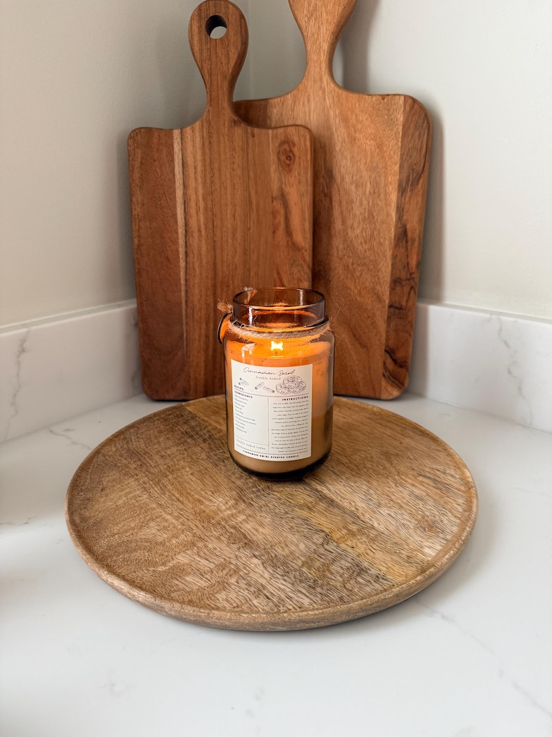 May include: A lit candle in a glass jar with a label sits on a wooden tray. Two wooden cutting boards stand behind the candle. The candle's flame glows, and the wooden items have a warm, natural color.