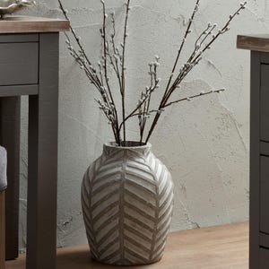 Pode incluir: Um vaso decorativo texturizado com um padrão de chevron em tons de cinza e branco. O vaso contém vários galhos de salgueiro. O vaso é um elemento decorativo para a casa.