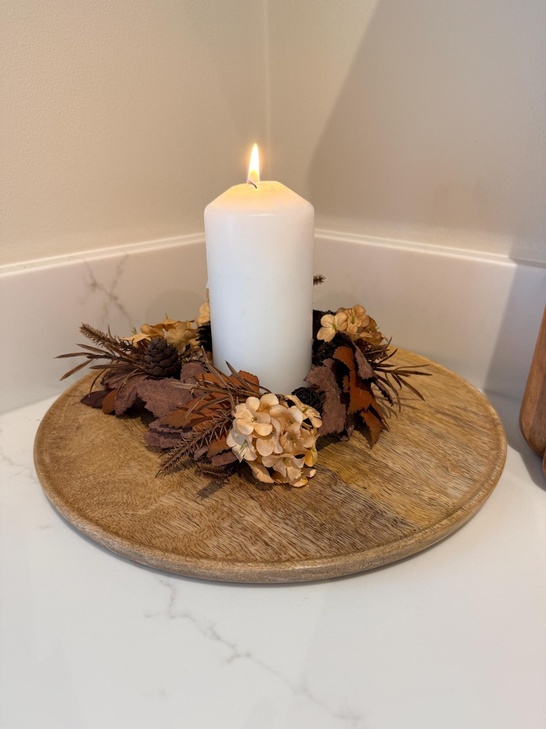 May include: A lit white pillar candle sits atop a round wooden tray, surrounded by a wreath of dried leaves, pinecones, and beige flowers. The candle flame glows brightly, creating a warm ambiance. The tray is placed on a white surface.