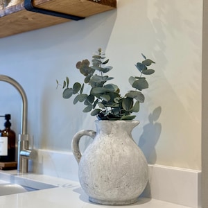 Op de afbeelding: Een witte, getextureerde keramische kan met een handvat, gevuld met groene eucalyptus takken. De kan staat op een wit aanrecht, met een kraan en plank op de achtergrond. Het thema is interieurdecoratie.