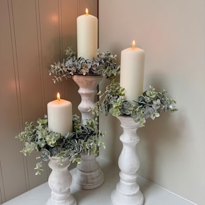 May include: Three white distressed candle holders with lit ivory candles and faux greenery. The candle holders are of varying heights and are placed on a white surface. The candles are cylindrical and have a warm glow.