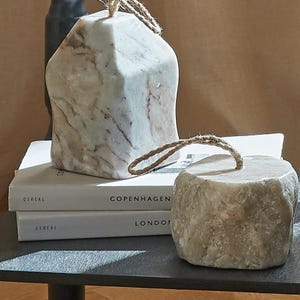 Peut inclure: Deux objets décoratifs en pierre avec des boucles en corde de jute pour suspendre. L'un a une forme de maison avec des veines blanches et brunes, l'autre est une pierre beige texturée et arrondie. Ils sont posés sur deux livres blancs.