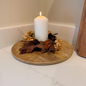 May include: A lit white pillar candle sits on a round wooden tray, surrounded by dried leaves, flowers, and pinecones. The tray is placed on a white surface, creating a warm, autumnal decorative display.