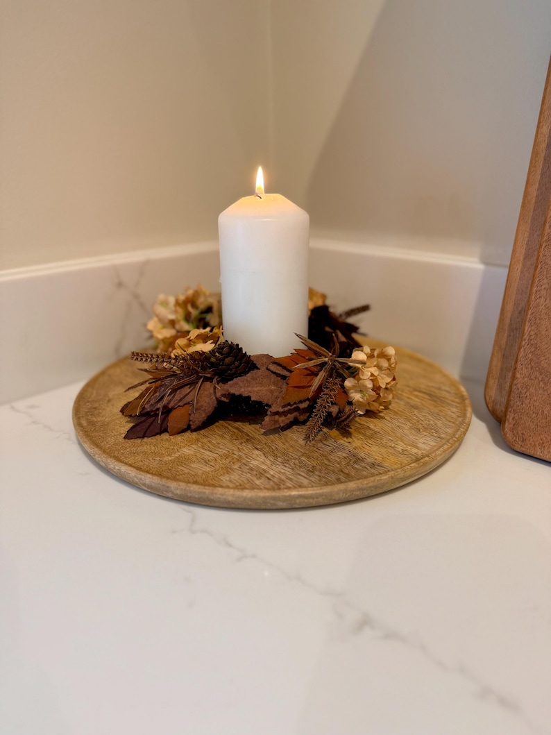 May include: A lit white pillar candle sits on a round wooden tray, surrounded by dried leaves, flowers, and pinecones. The tray is placed on a white surface, creating a warm, autumnal decorative display.