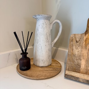 May include: A white ceramic pitcher with a handle and a textured surface sits on a round wooden tray. A small brown bottle with black reeds and the word "CALM" is also on the tray. A wooden cutting board is to the right.