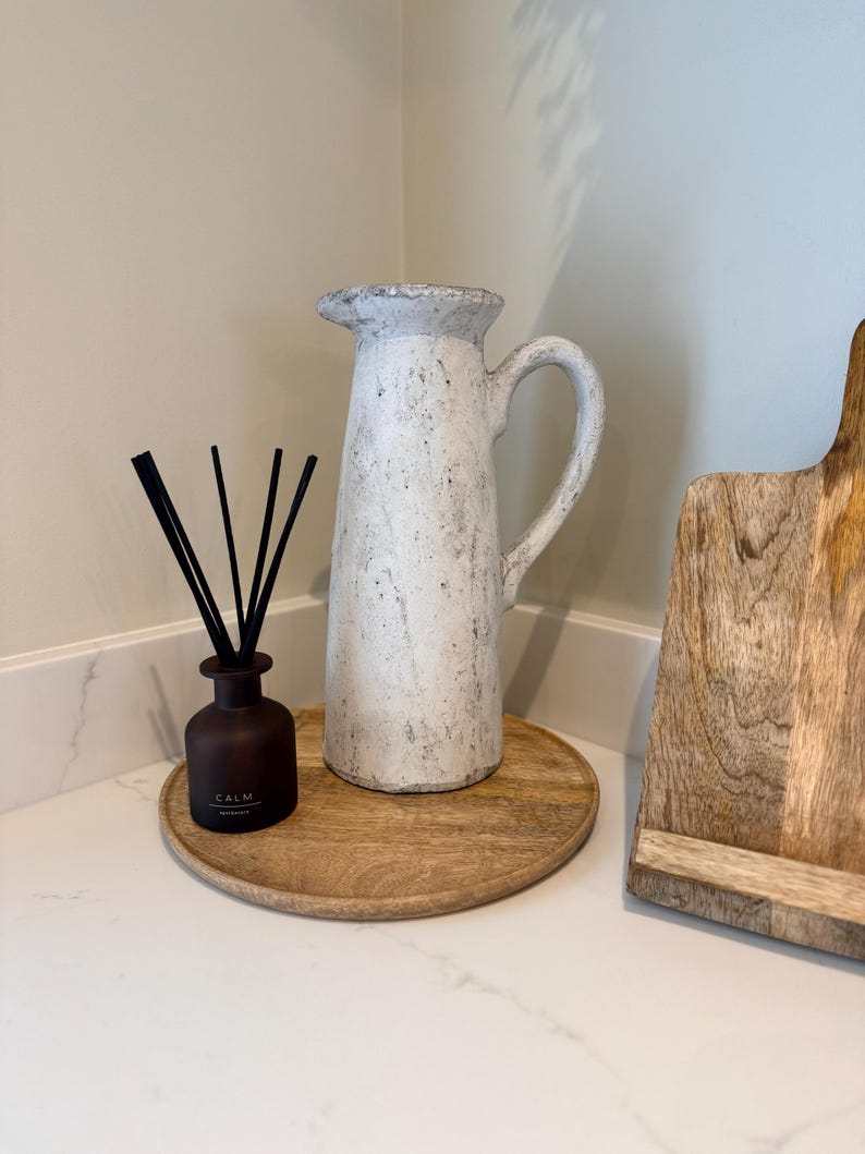 May include: A white ceramic pitcher with a handle and a textured surface sits on a round wooden tray. A small brown bottle with black reeds and the word "CALM" is also on the tray. A wooden cutting board is to the right.