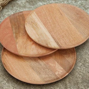May include: Three round wooden plates with a natural wood grain pattern. The plates are a warm, light brown color with variations in tone. The plates are stacked on top of each other, showcasing their circular shape and wooden texture.