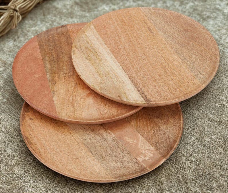May include: Three round wooden plates with a natural wood grain pattern. The plates are a warm, light brown color with variations in tone. The plates are stacked on top of each other, showcasing their circular shape and wooden texture.