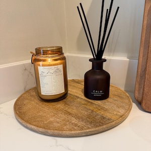 May include: A wooden tray holds a lit candle in a glass jar and a dark brown diffuser bottle with black reeds. The candle has a gold lid and a label. The diffuser bottle has the word "CALM" printed on it.