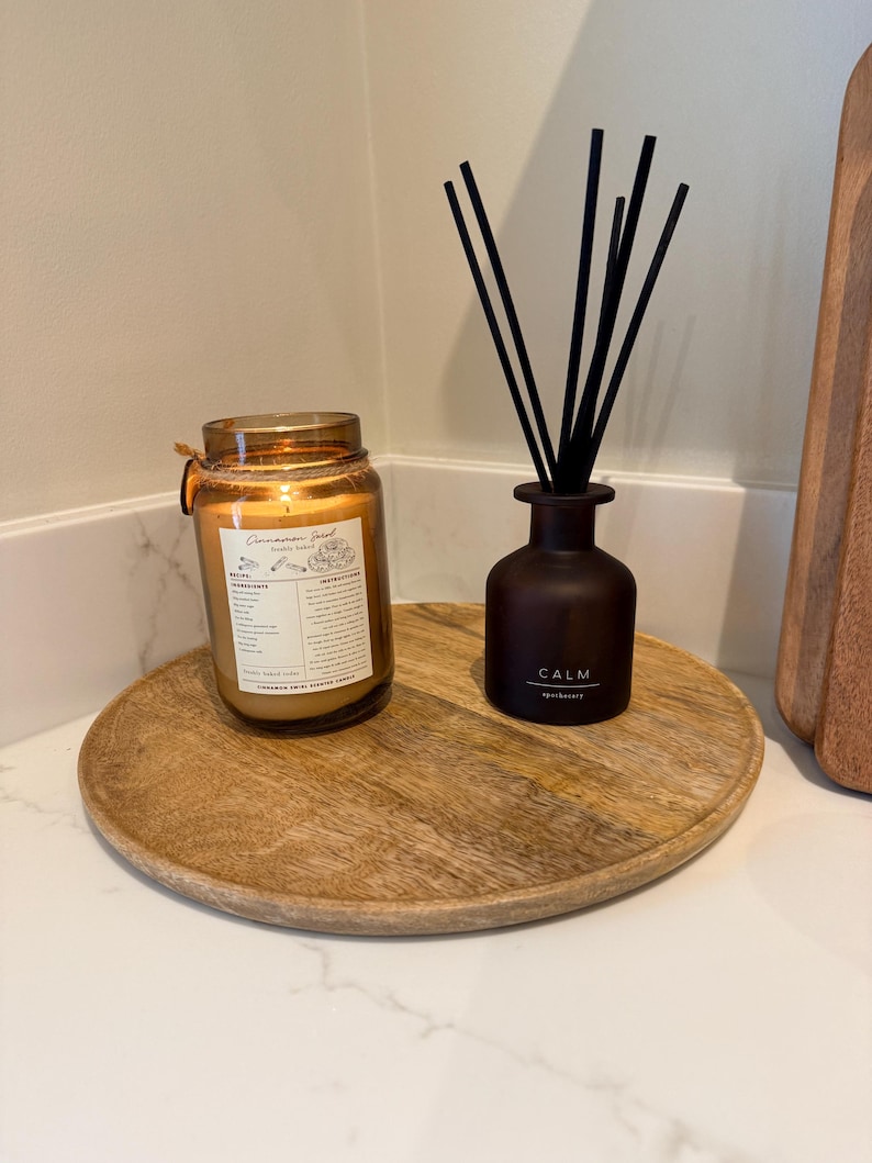 May include: A wooden tray holds a lit candle in a glass jar and a dark brown diffuser bottle with black reeds. The candle has a gold lid and a label. The diffuser bottle has the word "CALM" printed on it.