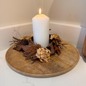 May include: A lit white pillar candle sits atop a round, wooden tray. The candle is surrounded by a decorative wreath of dried leaves, pinecones, and beige flowers. The tray is placed on a white surface.