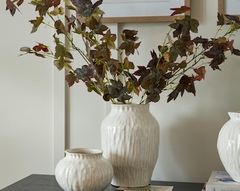 May include: Two white ceramic vases with textured vertical lines, one large and one small, sit on a dark surface. The vases hold leafy branches with green and burgundy leaves. The arrangement is a decorative home accent.