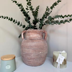 Puede incluir: Un jarrón grande y texturizado de color terracota con dos asas, lleno de ramas de eucalipto verdes. También hay una vela verde claro con una cinta blanca y un tarro verde claro con tapa de madera.