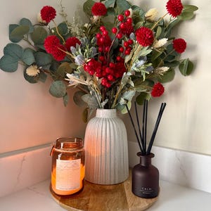 Könnte beinhalten: Eine dekorative Anordnung mit einem Strauß roter Blumen und Beeren in einer gerippten weißen Vase. Eine brennende Kerze in einem Glas und ein dunkelbrauner Diffusor mit schwarzen Stäbchen auf einem Holztablett.