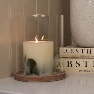 May include: A lit ivory pillar candle inside a glass cloche on a wooden base. The candle has a green and blue design. A textured white vase and two books with the words "AESTHETIC" and "ABSTRACT" are in the background.