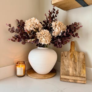 May include: A white textured vase with beige flowers and burgundy leaves. A wooden chopping board and a lit candle are on a white countertop. A wooden shelf is visible in the background.