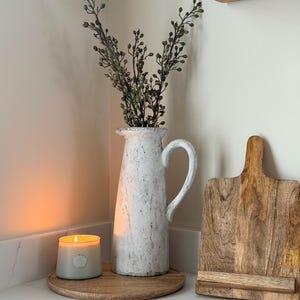 Op de afbeelding: Een witte, getextureerde kan met een handvat bevat bladrijke takken. Een brandende kaars in een grijze glazen pot en een houten snijplank zijn ook aanwezig. De items zijn gerangschikt op een houten dienblad, wat een gezellige, landelijke esthetiek creëert.