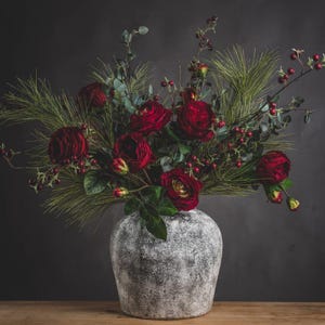 Op de afbeelding: Een bloemstuk met dieprode rozen, bessen en groen blad in een grijze vaas met textuur. Het boeket bevat dennenachtige elementen en eucalyptusbladeren. De vaas staat op een houten oppervlak tegen een donkergrijze achtergrond.