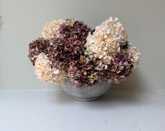 Könnte beinhalten: Ein Arrangement aus künstlichen Hortensien in einer Betonschale. Die Blumen sind eine Mischung aus Farben, darunter hellrosa, creme und tiefes Burgunderrot. Die Schale ist rund und steht auf einer weißen Arbeitsplatte, was eine neutrale und elegante Präsentation ergibt.