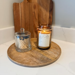 May include: A wooden serving tray holds a lit candle in a glass jar, a glass jar with matches, and two wooden cutting boards. The candle has a label with text. The items are arranged on a white countertop.