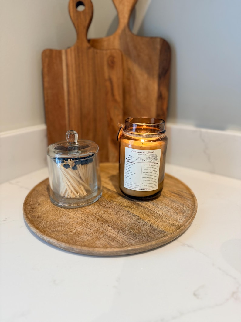 May include: A wooden serving tray holds a lit candle in a glass jar, a glass jar with matches, and two wooden cutting boards. The candle has a label with text. The items are arranged on a white countertop.