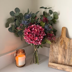 Puede incluir: Un ramo de flores artificiales con una gran hortensia rosa vibrante, cardo azul y acentos burdeos, con hojas de eucalipto verdes. También hay una vela encendida en un tarro de cristal y una tabla de cortar de madera.