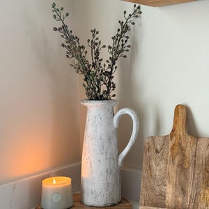 May include: A white textured pitcher with a handle holds greenery. A lit candle sits on a wooden tray, next to a wooden cutting board. A wooden shelf is in the background. The scene has a rustic, farmhouse aesthetic.