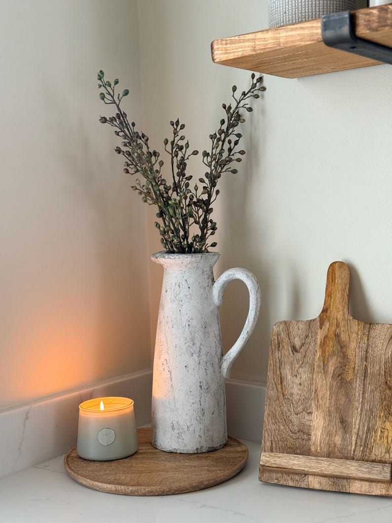 May include: A white textured pitcher with a handle holds greenery. A lit candle sits on a wooden tray, next to a wooden cutting board. A wooden shelf is in the background. The scene has a rustic, farmhouse aesthetic.