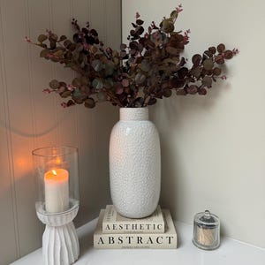 Peut inclure: Un vase blanc texturé rempli de branches d'eucalyptus vert foncé et bordeaux est posé sur deux livres intitulés "Aesthetic" et "Abstract". Une bougie allumée dans un photophore en verre et un petit pot en verre sont également sur la surface blanche.