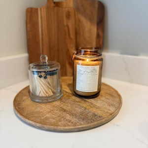 May include: A wooden serving tray holds a lit candle in a glass jar, a glass jar of matches, and two wooden cutting boards. The candle label reads "Cinnamon Jar". The tray and cutting boards are made of wood.
