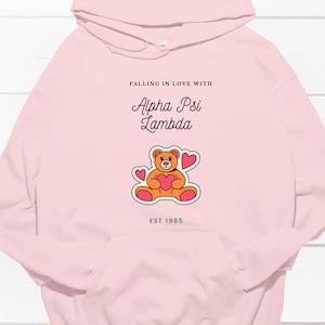 Op de afbeelding: Roze hoodie met een afbeelding van een teddybeer die een hart vasthoudt en de tekst "Falling in love with Alpha Psi Lambda Est 1985".