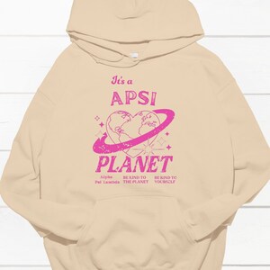 Könnte beinhalten: Ein beiger Hoodie mit einem rosafarbenen Grafikdruck, der "It's a APSI PLANET" mit einer herzförmigen Erde und einem Ring darum herum sagt. Der Text unter der Grafik lautet "Alpha Pal Lambda BE KIND TO THE PLANET BE KIND TO YOURSELF".