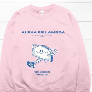 Könnte beinhalten: Rosafarbener Pullover mit dem Text "ALPHA PSI LAMBDA" und "ANNUAL CARNE ASADA 2023" in blauer Schrift. Ein Cartoon-Taco mit Beinen und Armen hält einen Teller mit einem lächelnden Gesicht. Unter dem Taco steht der Text "MIAMI UNIVERSITY OXFORD, OH" in blauer Schrift.