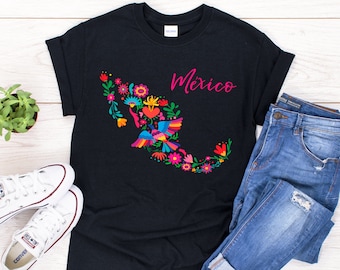 May include: Black t-shirt featuring a vibrant floral and bird design in the shape of Mexico. The word "Mexico" is written in pink script. The shirt is styled with blue jeans and white trainers.
