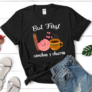 May include: Black t-shirt with white text that reads "But First Conchas y Churros". The text is accompanied by a cartoon image of a concha pastry and a cup of coffee.