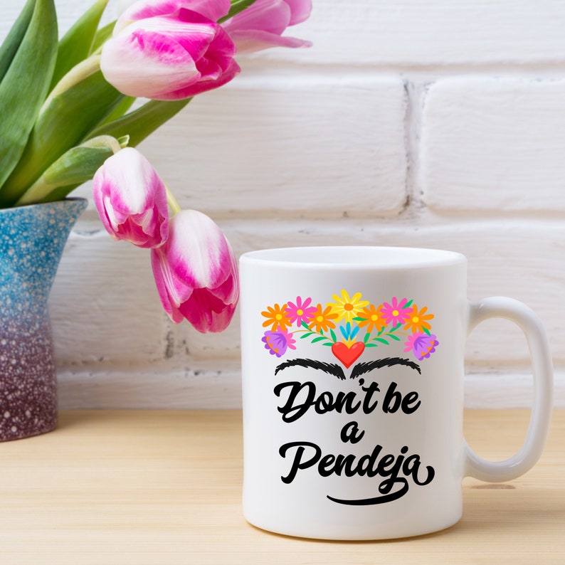 May include: White ceramic mug with a black and white graphic that says "Don't be a Pendeja" with a colorful floral design above the text. The mug has a handle on the right side.