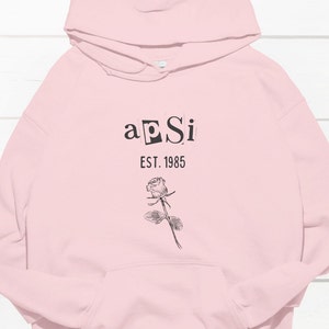 Op de afbeelding: Roze hoodie met een zwarte afbeelding met de tekst "a pSi EST. 1985" en een zwart-witte roos illustratie.