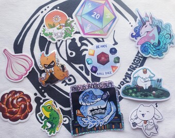 May include: A collection of colorful stickers featuring various designs. Included are a d20 die, a unicorn, a frog, a cat-like character, a garlic bulb, a flower, a dragon reading a book with the text "PROUD BOOKWYRM", and other whimsical illustrations.