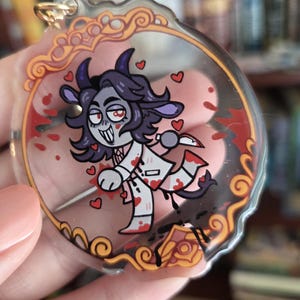 Op de afbeelding: Een heldere acryl bedel met een cartoon illustratie van een geitachtig karakter met een mes, omringd door harten en bloedspetters. De bedel heeft een goudkleurige ring voor bevestiging en een sierlijke rand.