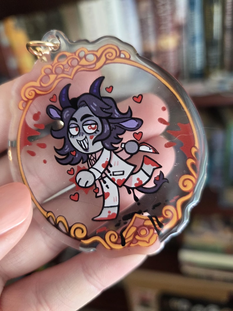Op de afbeelding: Een heldere acryl bedel met een cartoon illustratie van een geitachtig karakter met een mes, omringd door harten en bloedspetters. De bedel heeft een goudkleurige ring voor bevestiging en een sierlijke rand.