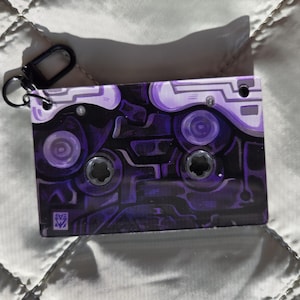 May include: A rectangular keychain shaped like a cassette tape featuring a dark purple and black abstract circuit board pattern. The design includes two circular cutouts, a small black metal lobster clasp attached to the top left corner, and a small square logo in the bottom left corner.