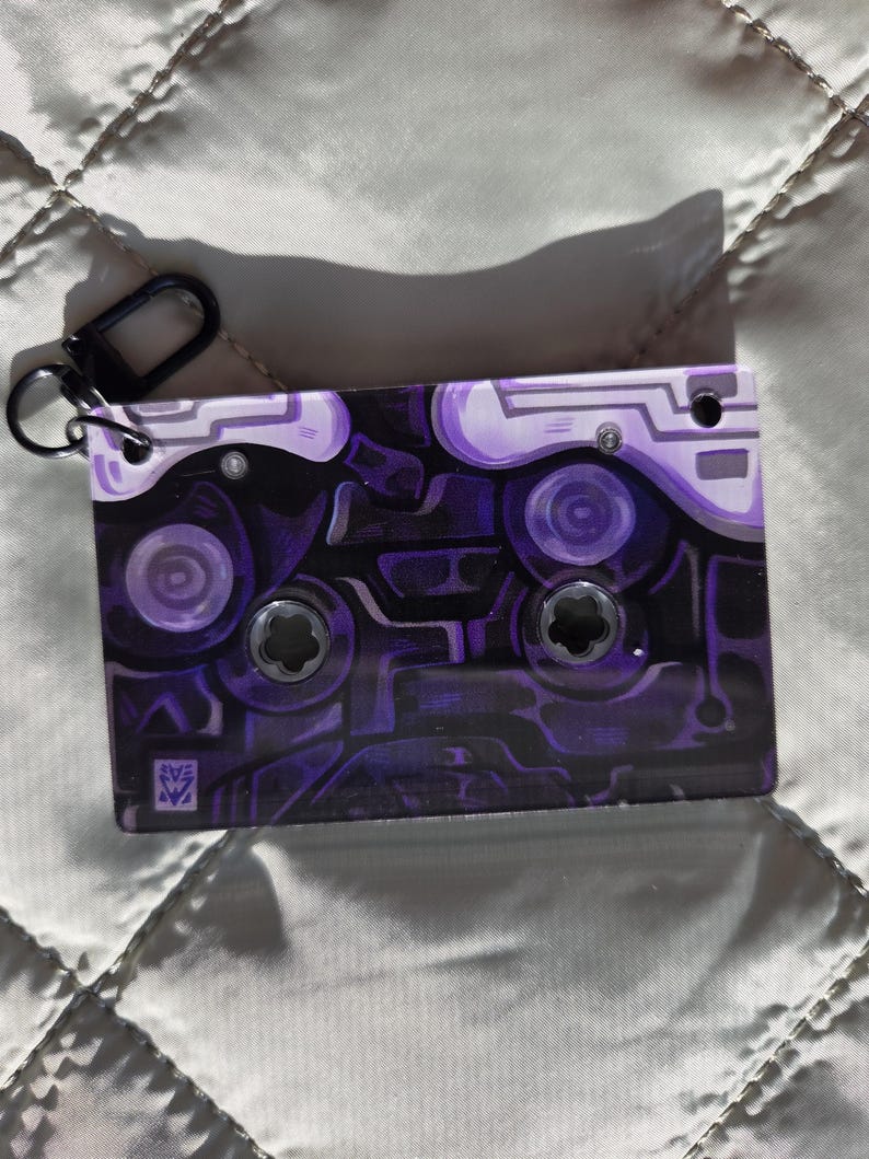 May include: A rectangular keychain shaped like a cassette tape featuring a dark purple and black abstract circuit board pattern. The design includes two circular cutouts, a small black metal lobster clasp attached to the top left corner, and a small square logo in the bottom left corner.