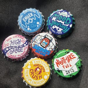May include: A collection of six colorful bottle caps with various designs and text. The caps feature phrases like "Nightmare Fuel 2012" and "Mood Whiplash". The caps are on a dark gray surface.