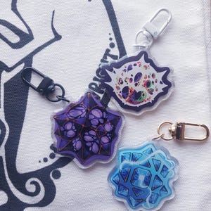 May include: Four colorful acrylic keychains with lobster claw clasps. The keychains feature unique, geometric designs in purple, blue, and a multi-colored skull-like pattern. The keychains are displayed on a white fabric surface.
