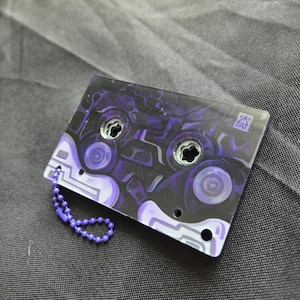 May include: A rectangular acrylic keychain shaped like a cassette tape featuring a dark purple and black abstract pattern. The design includes two circular cutouts, a small logo in the corner, and a purple ball chain attached to the side.